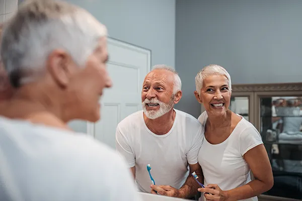 Elderly white couple smiling and brushing their teeth in the bathroom mirror as instructed by Surf City Oral and Maxillofacial Surgery in Huntington Beach, CA